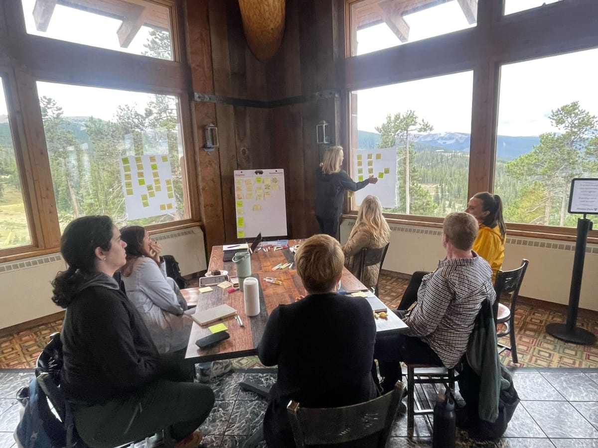 Proof Over Promises attendees in a working group session at Winter Park, clustering sticky notes on flip charts in front of a wall of mountain windows
